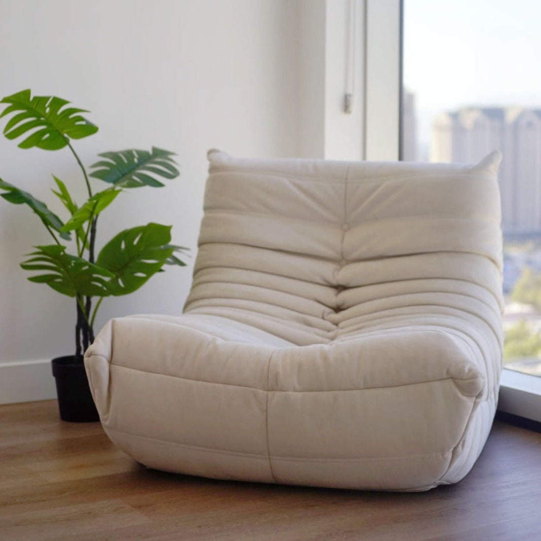A soft, white Chillax Lounger Sofa Chair sits in a corner by large windows beside a green potted plant on a wooden floor, offering a luxurious lounging experience.