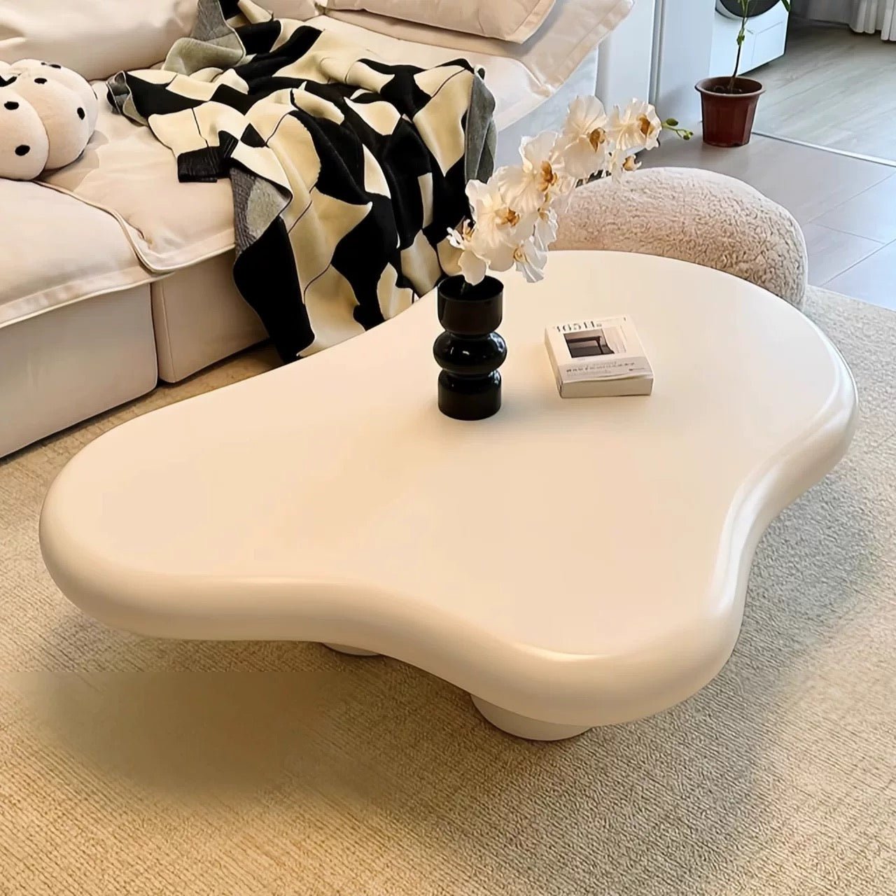 A modern living room features the Modern Cloud-Like Coffee Table with an eco-friendly white fiberglass finish. A vase of white flowers and a book sit atop it, while a black and white patterned blanket drapes over the sofa, embodying minimalist aesthetics.