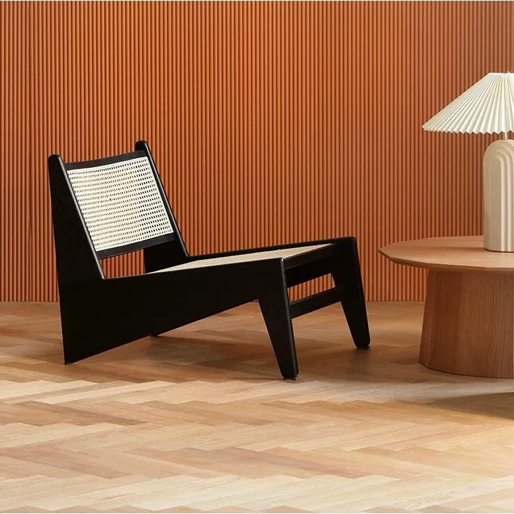 A 袋鼠椅 with a woven black back and seat sits beside a round wooden table topped with a white lamp, set against a ribbed orange wall and light wood floor.