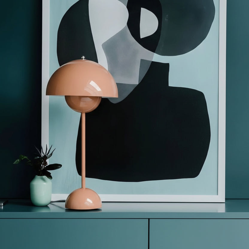 A Large Bud Table Lamp | Plug-In Edition with a cast iron base and bud-shaped peach shade, plus a small plant in a light green pot, sit atop a teal cabinet before an abstract black and gray artwork.