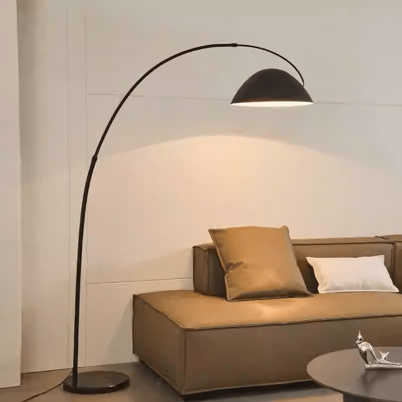 A modern mid-century Arched Floor Lamp in Matte Black stands next to a beige sofa, adorned with a pillow and a cushion.
