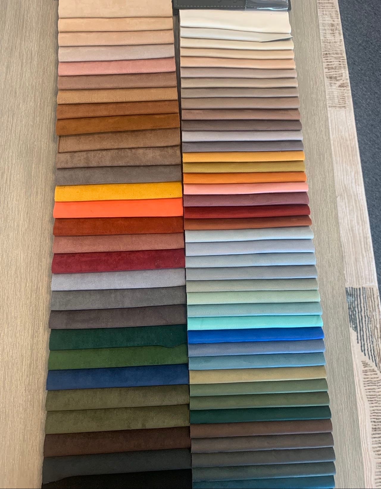 A table showcases neatly arranged fabric swatches in diverse colors, such as brown, red, orange, yellow, green, blue, and gray—perfectly complementing the luxurious experience on the Large Chillax Lounger – 2 Seat Sofa.