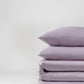 A neatly stacked pile of luxury bedding in light purple, featuring two pillows and the Luxe Tencel-Cotton Fitted Sheet, stands against a plain white background.