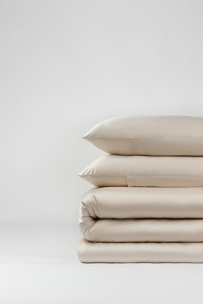 A stack of neatly folded beige luxury bedding, including eco-friendly pillowcases and the Luxe Tencel-Cotton Fitted Sheet, is displayed against a plain white background.