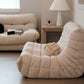 A cozy living room features a Large Chillax Lounger – 2 Seat Sofa, a plush beige sofa, a wooden side table with a table lamp, and a decorative knot cushion. Blinds on the window let light gently filter through the room.
