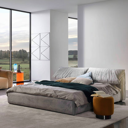 A modern bedroom showcases a sleek Grey Velvet Upholstered Bed Frame with gray and teal bedding, complemented by a plush mustard fabric ottoman. A curved floor lamp and abstract wall art add elegance, while a large window frames the scenic landscape view.
