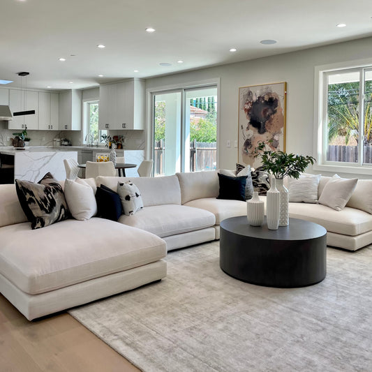 Spacious modern living room with a Snowdrift L-Shaped Sectional Sofa, dark round coffee table, abstract wall art, and large windows. Kitchen with island visible in the background.