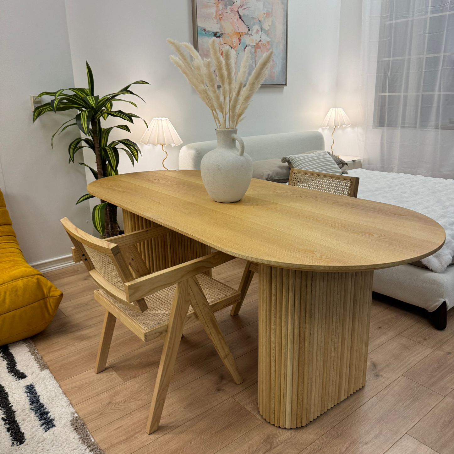An Oval Shaped Solid Wood Dining Table with an eco-friendly lacquer finish holds a vase of dried pampas grass. Nearby, two chairs, a plant, and a floor lamp complement the setting. In the background, a white sofa and wall art piece enhance the harmonious look.
