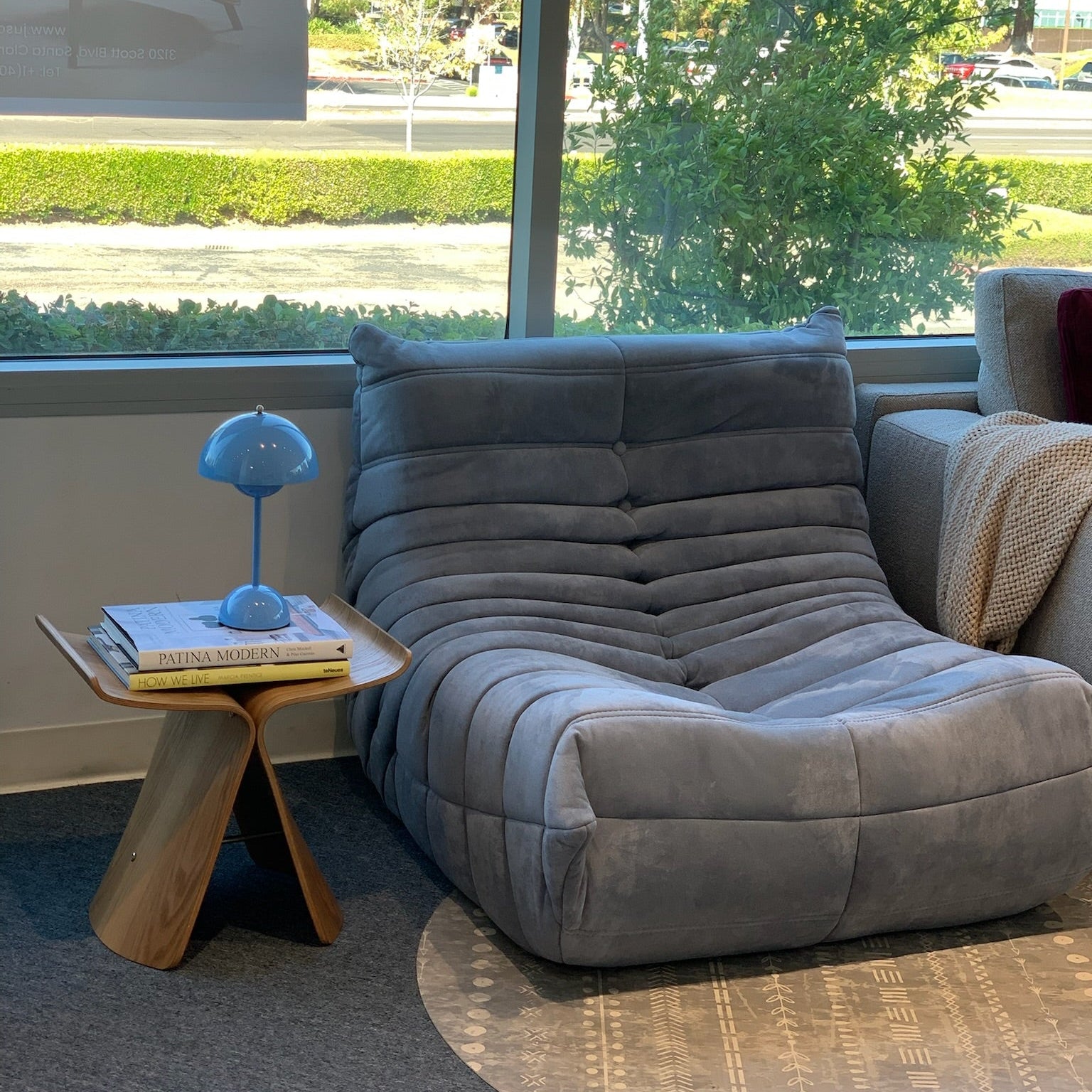 Gray-blue sofa in a room with a window view of trees and a small table with a lamp and books.