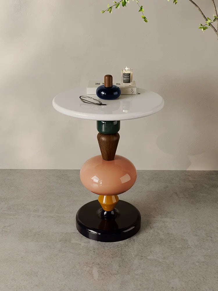 The 糖葫芦茶几 is a round white side table with a multicolored geometric base. It displays a blue vase, lit candle, and eyeglasses, set against a neutral wall and floor.
