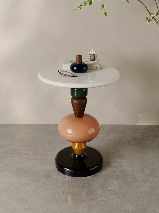The 糖葫芦茶几 is a round white side table with a multicolored geometric base. It displays a blue vase, lit candle, and eyeglasses, set against a neutral wall and floor.
