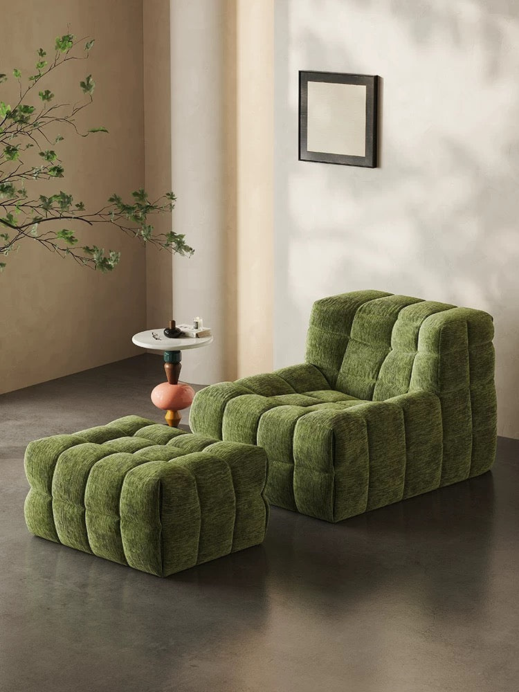 A green 休闲椅2 with matching ottoman sits on a concrete floor next to a small white side table and a leafy branch. A framed art piece hangs on the wall.
