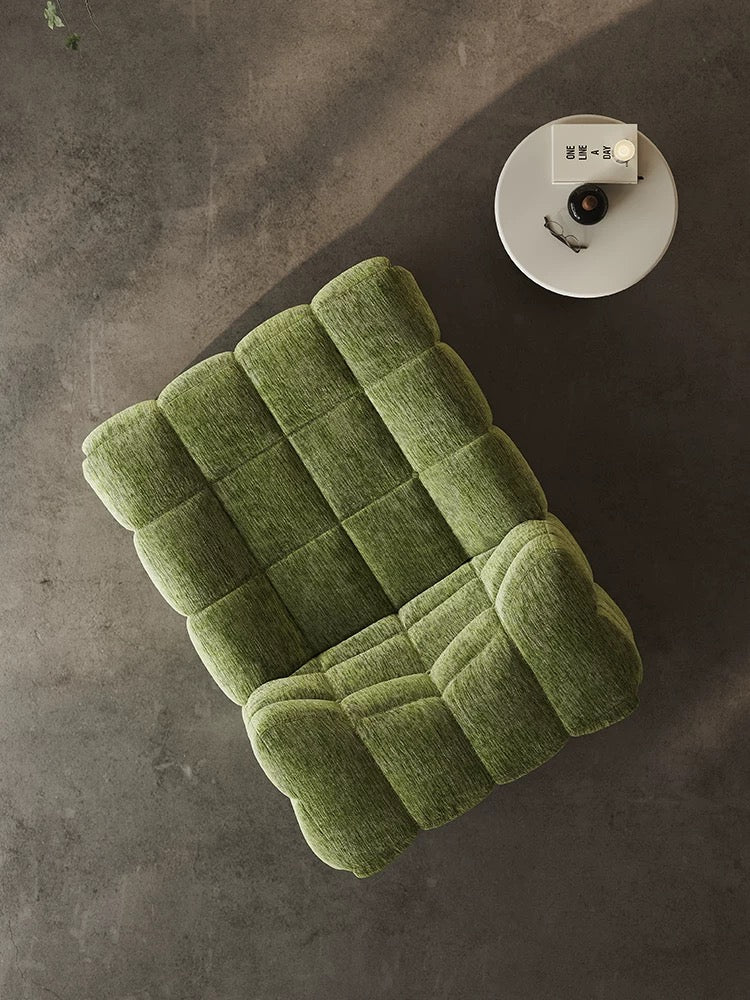 The 休闲椅2, a green cushioned armchair, is placed on a concrete floor beside a round white side table holding a book and coffee cup.