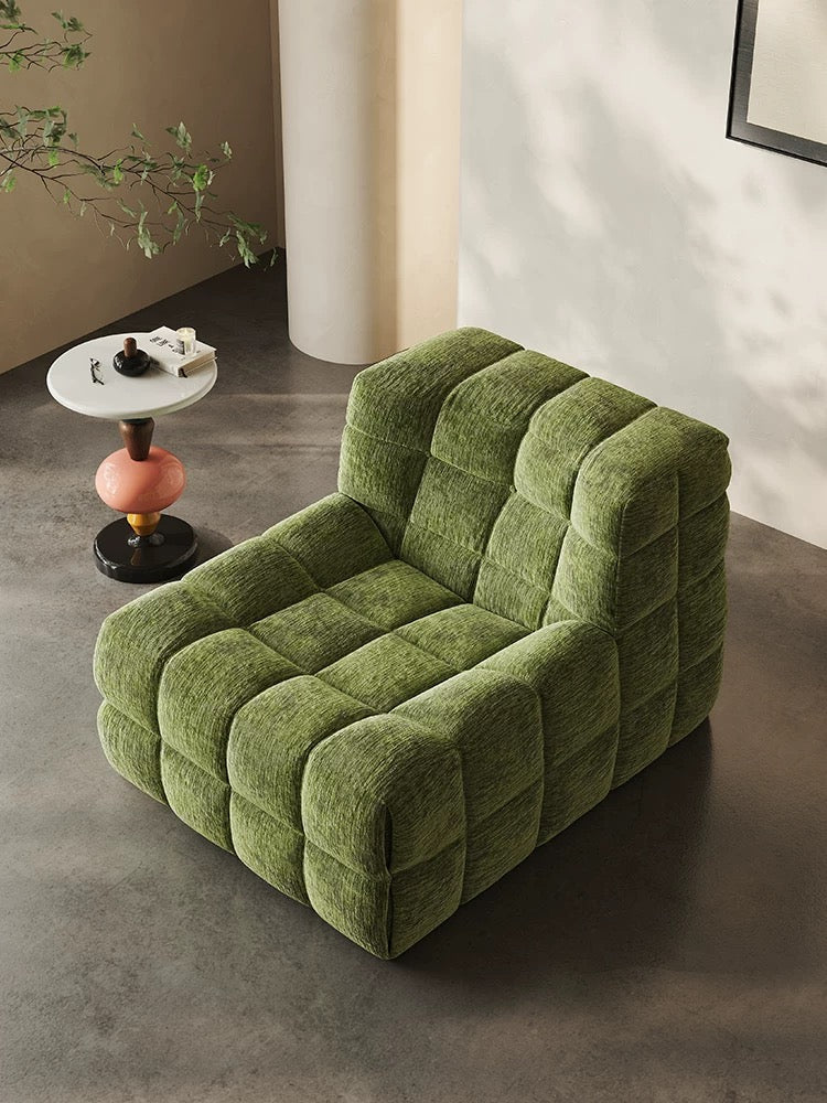 A green, plush, modular armchair (休闲椅2) with a grid pattern sits next to a small round side table holding a cup, saucer, and decor on a gray floor in a minimalist modern room.