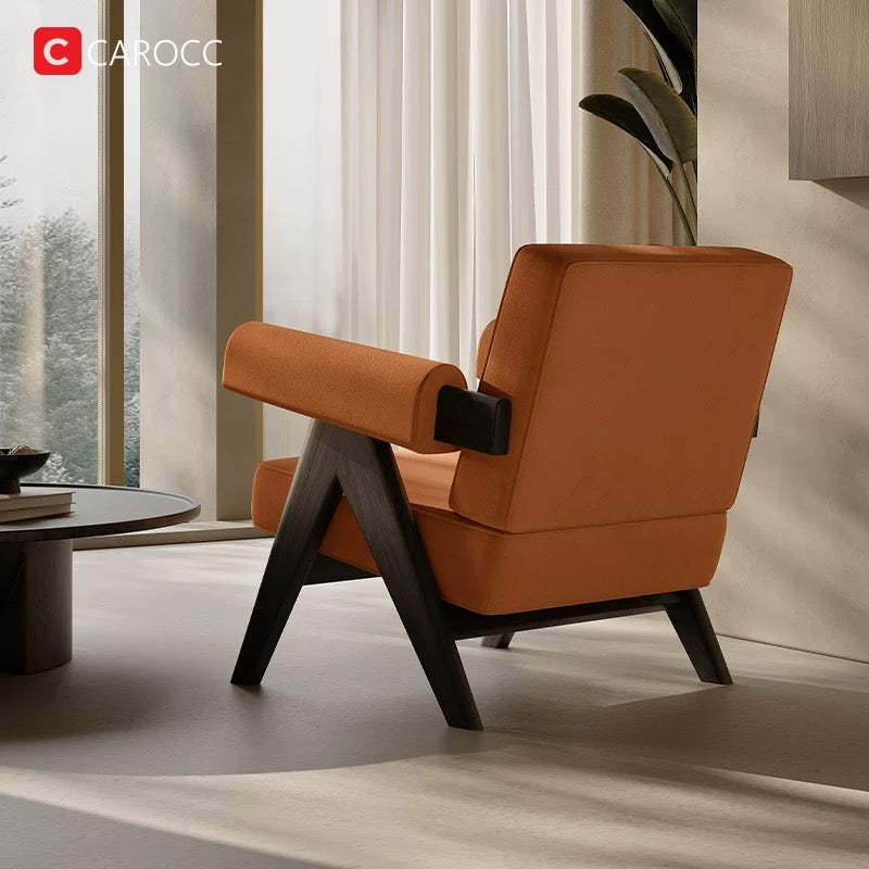 A 休闲椅 with orange upholstery and black wooden legs is placed in a modern living room by large windows and a round table, sunlight streaming in.