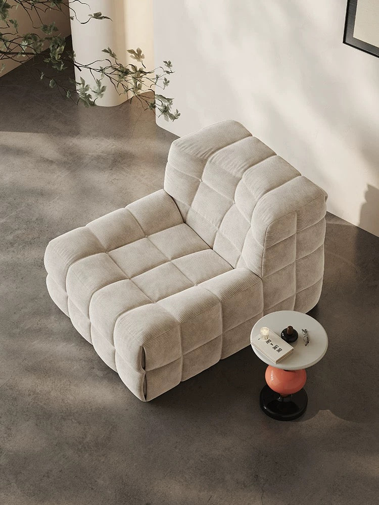 A modern beige 休闲椅2 sits on a concrete floor beside a small round side table holding a remote, cup, and snack plate.