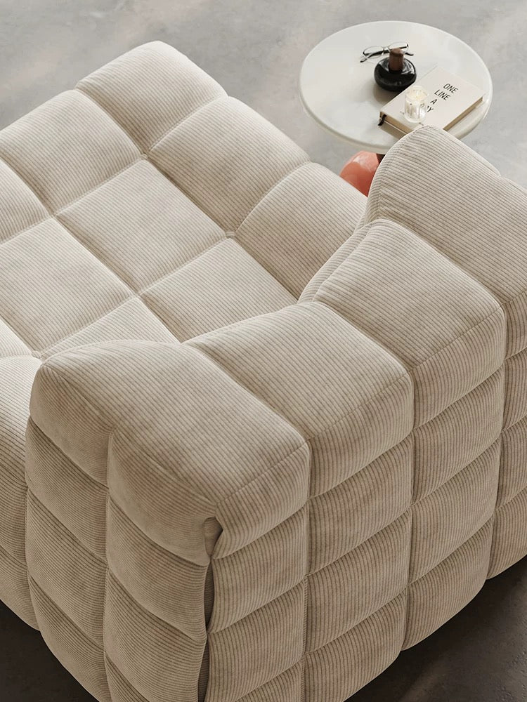 A beige corduroy grid-patterned 休闲椅2 is placed next to a round white side table topped with a glass, a book, and a small dark jar.