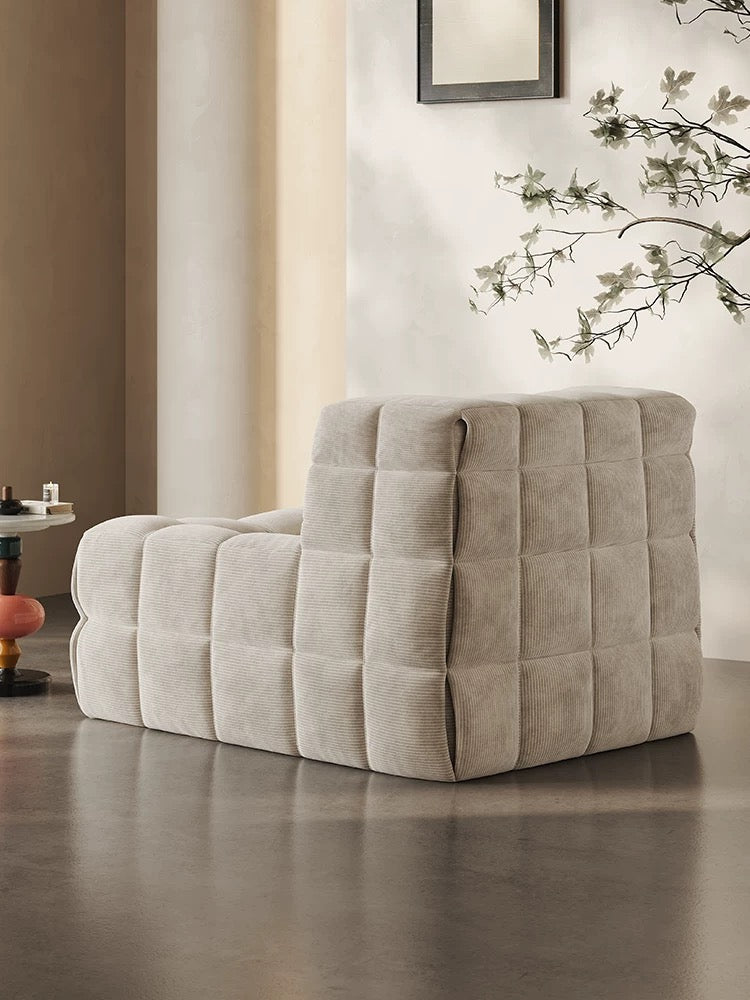 The 休闲椅2, a modern light beige tufted lounge chair, sits on a polished concrete floor in a minimalist room with neutral walls and a tall plant in the background.
