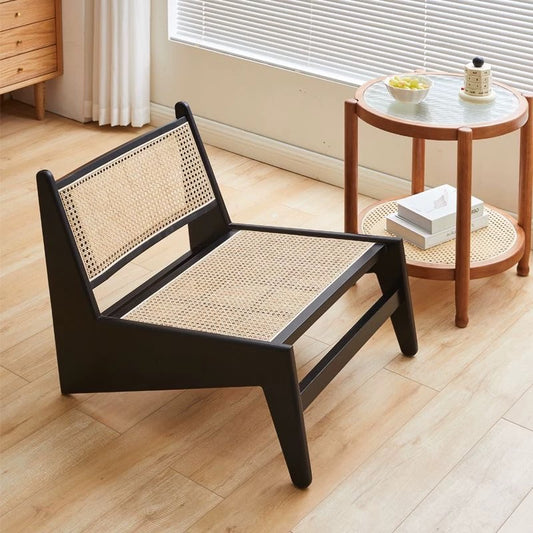 The 袋鼠椅, a low modern chair with black frame and woven rattan seat and backrest, stands beside a round wooden side table in a sunlit room with light wood floors.
