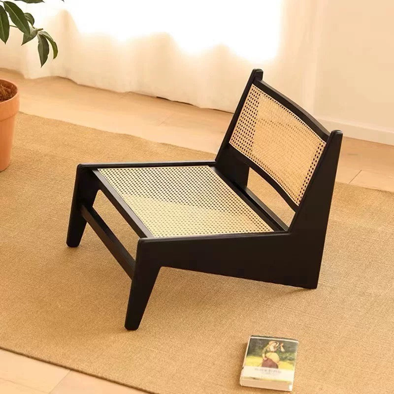 The 袋鼠椅, a low wooden chair with a woven cane seat and backrest, is placed on a beige rug beside a potted plant and a book on the floor.