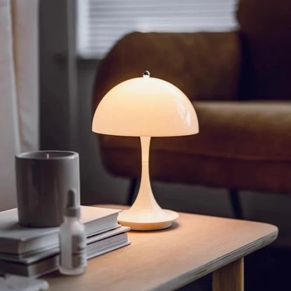 The Cordless Mashroom Lamp, a modern rechargeable table lamp with a dome-shaped shade, sits on a wooden table beside books, a mug, and a dropper bottle; in the background are blinds and a brown chair.