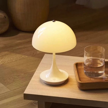 The Cordless Mashroom Lamp, a compact modern white rechargeable light, glows warmly beside a glass of water and a wooden tray on a light wooden table.