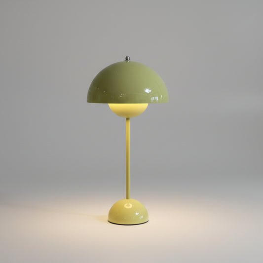 A Large Bud Table Lamp | Plug-In Edition with Cast Iron Base featuring a yellow bud shade glows beside a blue and white ceramic jar on a wooden surface against a light-colored wall.