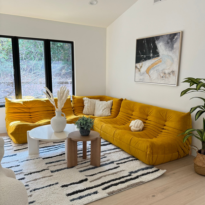 A cozy living room showcases a Large Chillax Lounger in mustard yellow with buttery-soft upholstery, black-framed windows, abstract wall art, a striped rug, and a small round white table decorated with accents.
