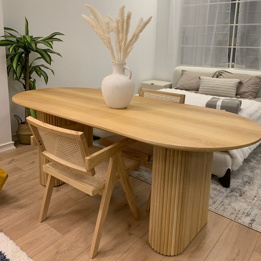 A minimalist room features an Oval Shaped Solid Wood Dining Table topped with premium sintered stone and two chairs, a white vase with pampas grass, and a bed with pillows in the background, complemented by a potted plant in the corner.
