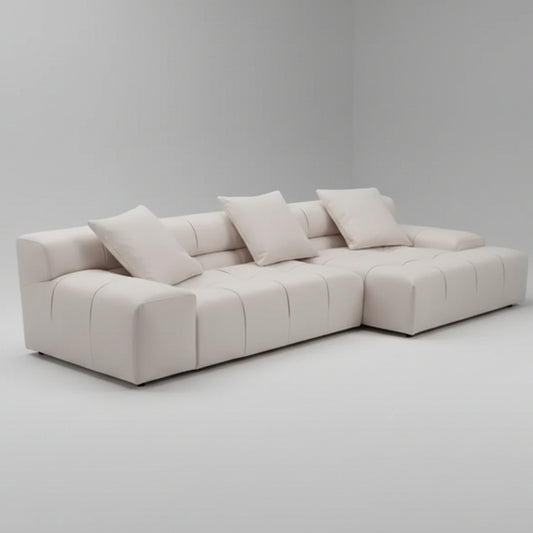 The Verve Tufted Modular Sofa with Chaise in beige is a luxurious, modern L-shaped design, featuring four matching cushions against its pristine white backdrop.