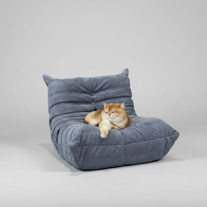A fluffy cat lounges on a soft, grey Chillax Lounger Sofa Chair made with high-density foam in a cozy living room. Nearby are a blue armchair, a small table, and a toy kitchen set.