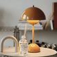 A round orange Cordless Bud Lamp with adjustable brightness sits beside a "Bois de Santal" glass reed diffuser on a white surface, with shelves and books softly blurred in the background.